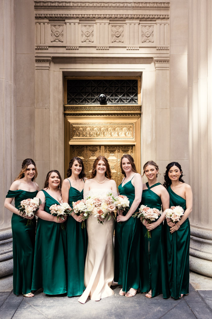 Portrait of bride in a white dress with six bridesmaids in green dresses, all holding bouquets, standing in front of a golden door flanked by marble columns in downtown Chicago