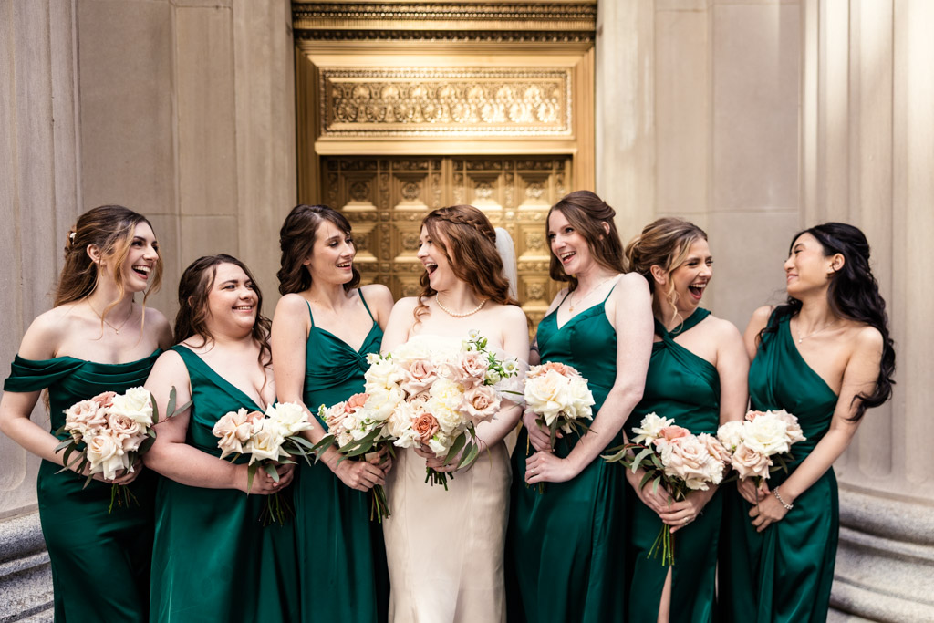 Candid photo of bride and bridesmaids in green dresses smiling and laughing together while holding bouquets in front of a golden door flanked by marble columsn in downtown Chicago