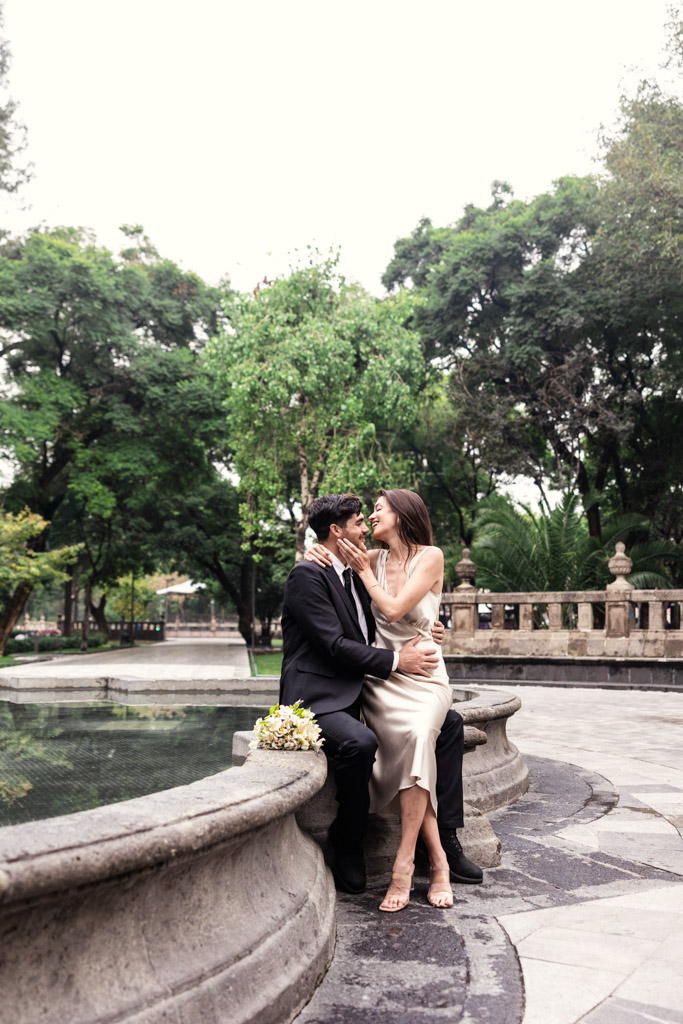 A couple dressed formally sits on a stone fountain, embracing and smiling, surrounded by lush greenery—capturing the romance of a Mexico City elopement.