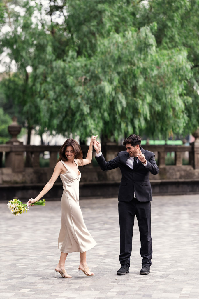 A joyful couple dressed up, holding hands and dancing outdoors during their Mexico City elopement; the woman holds a bouquet of flowers.