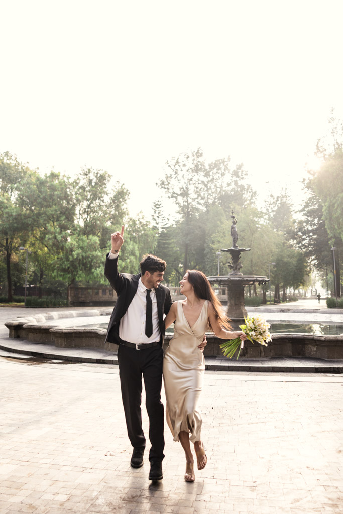 A joyful couple, dressed formally, walks arm in arm near a fountain in Alameda Central, holding flowers and smiling during their Mexico City elopement