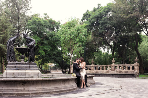 A couple holds each other seated on the edge of a stone fountain in lush Alameda Central during their Mexico City elopement