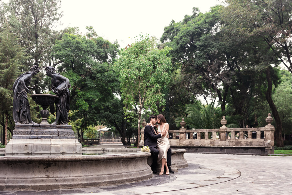 A couple holds each other seated on the edge of a stone fountain in lush Alameda Central during their Mexico City elopement