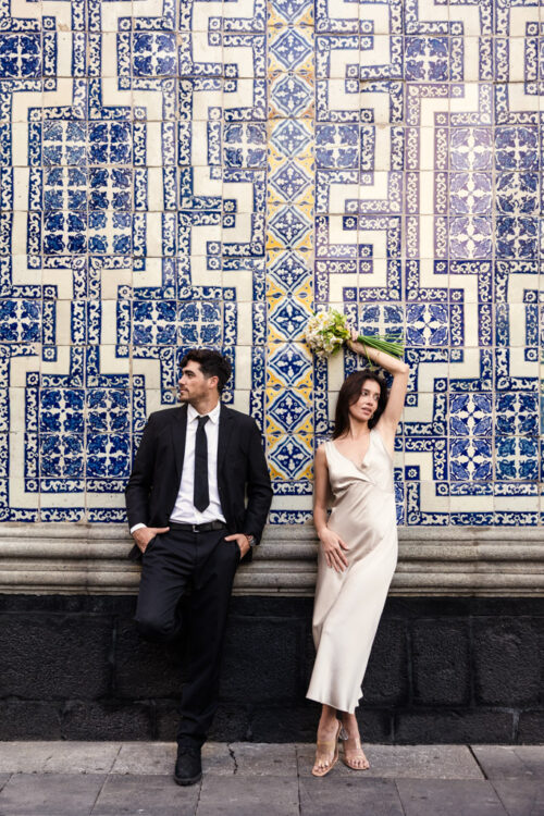 A couple in formal attire poses against a decorative blue and white tiled wall at La Casa de los Azulejos, the woman holding a bouquet during their Mexico City elopement