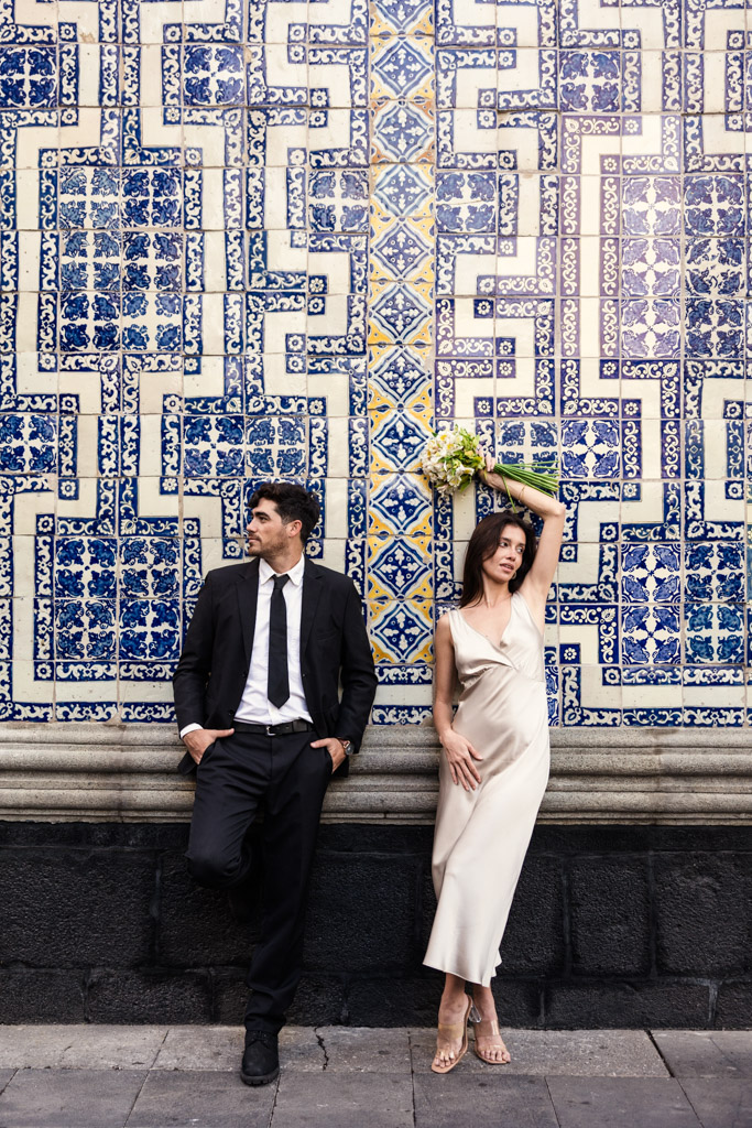 A couple in formal attire poses against a decorative blue and white tiled wall at La Casa de los Azulejos, the woman holding a bouquet during their Mexico City elopement