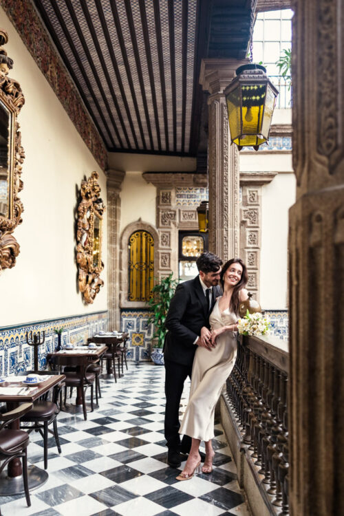 A couple dressed formally smiles and embraces on a balcony in La Casa de los Azulejos with checkered floors during their Mexico City elopement
