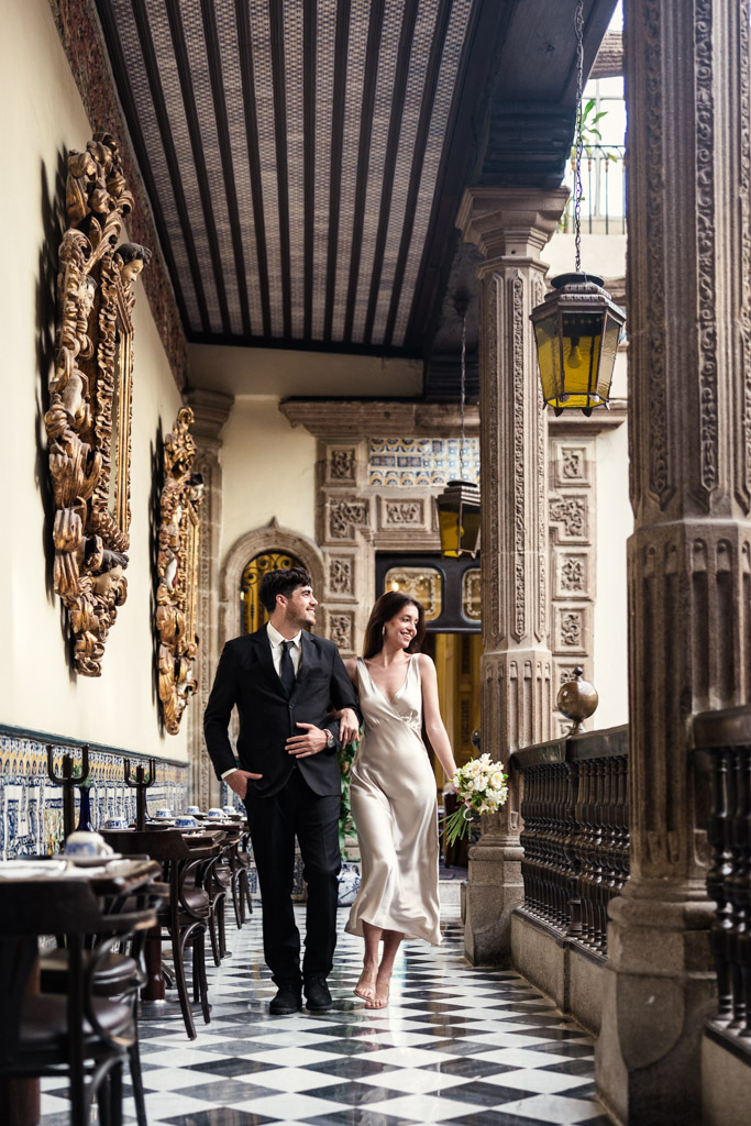 A couple in formal attire walks happily along a grand, ornately decorated balcony with checkered floors at La Casa de los Azulejos during their Mexico City elopement