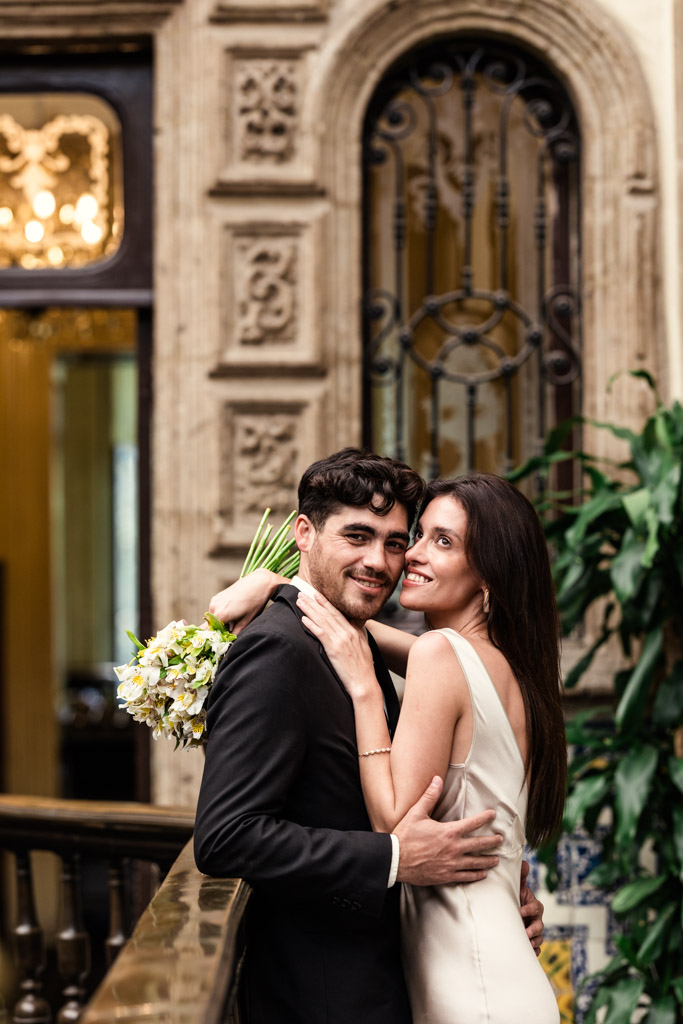 A smiling couple in formal attire embrace on a balcony at La Casa de los Azulejos during their Mexico City elopement, the woman holding a bouquet of flowers