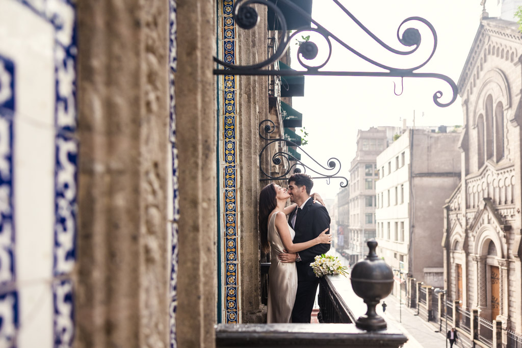 A couple embraces on a city balcony decorated with blue tile at La Casa de los Azulejos during their Mexico City elopement with city buildings in the background