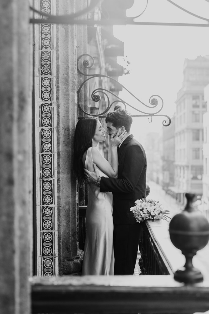 Romantic black and white photo of couple on a La Casa de los Azulejos balcony, smiling at each other during their Mexico City elopement, with city buildings in the background