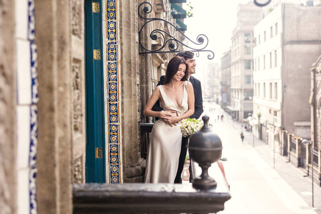 Happy couple in wedding attire stands on a balcony at La Casa de los Azulejos, smiling and holding each other during their Mexico City elopement