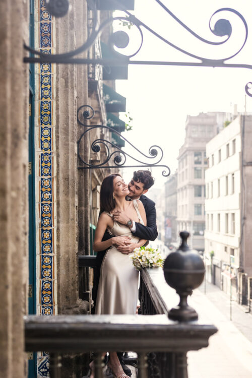 A couple embraces and smiles on a La Casa de los Azulejos balcony with ornate ironwork and buildings in the background during their Mexico City elopement
