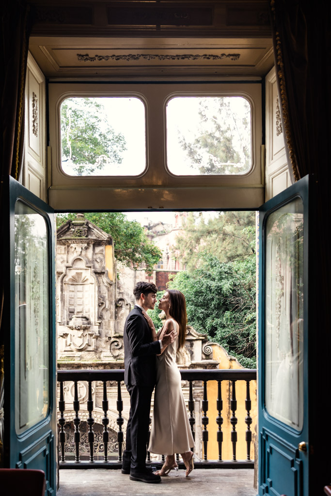 Couple stands close together on a balcony at La Casa de los Azulejos during their Mexico City elopement, framed by open doors, with historic stonework and trees behind them