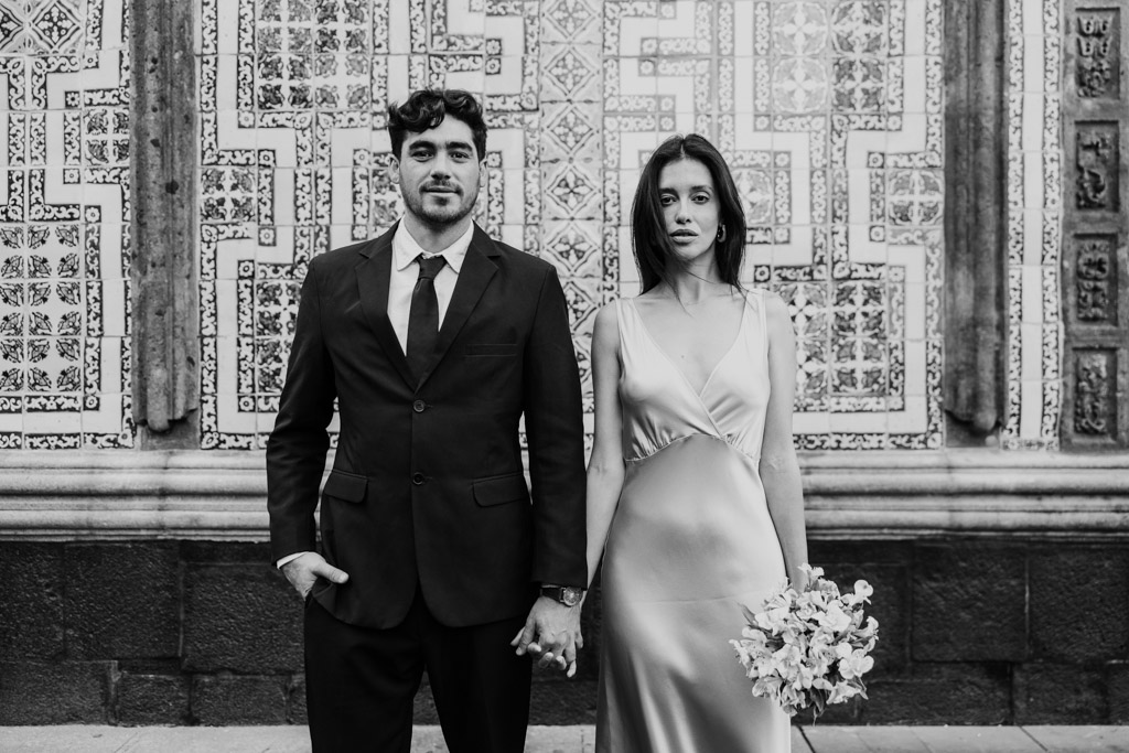 Black and white portrait of newlywed couple in formal attire holding hands, standing in front of an ornate patterned wall at La Casa de los Azulejos during their Mexico City elopement