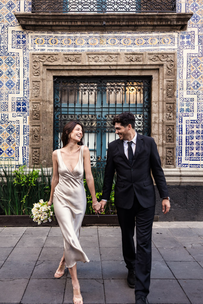 A smiling couple in formal attire holds hands and walks in front of the blue tiled facade of La Casa de los Azulejos during their Mexico City elopement
