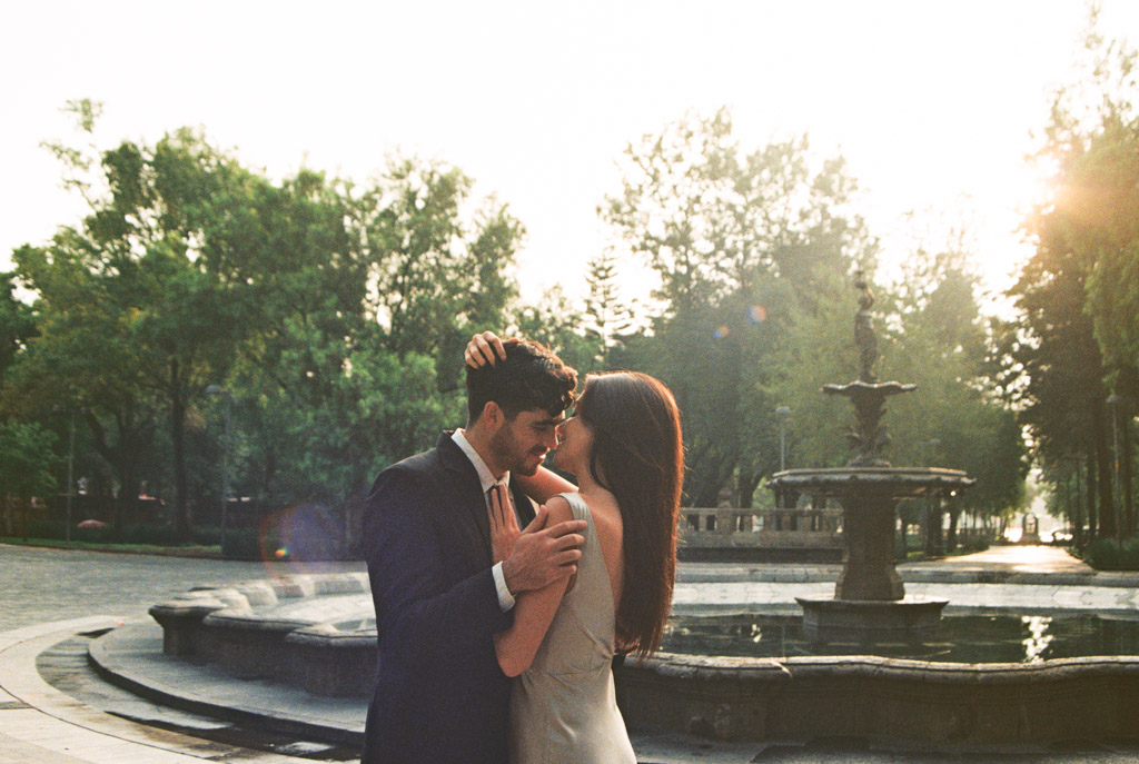 Film photo of couple holding each other beside a fountain in sunlit, tree-lined Alameda Central during their Mexico City elopement