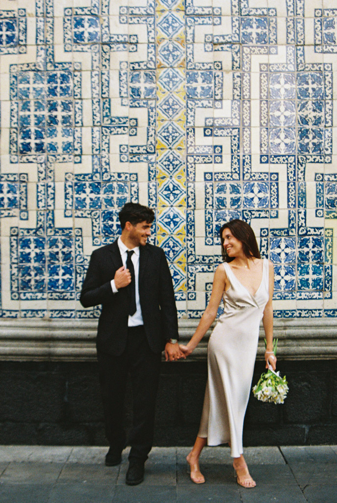 A couple in formal attire holds hands and smiles in front of a blue and white patterned tile wall, capturing the joy of their Mexico City elopement.