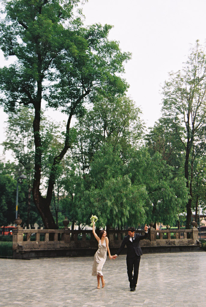 Film photo of couple in formal attire joyfully walking hand-in-hand on a stone path in Alameda Central beside tall green trees during Mexico City elopement