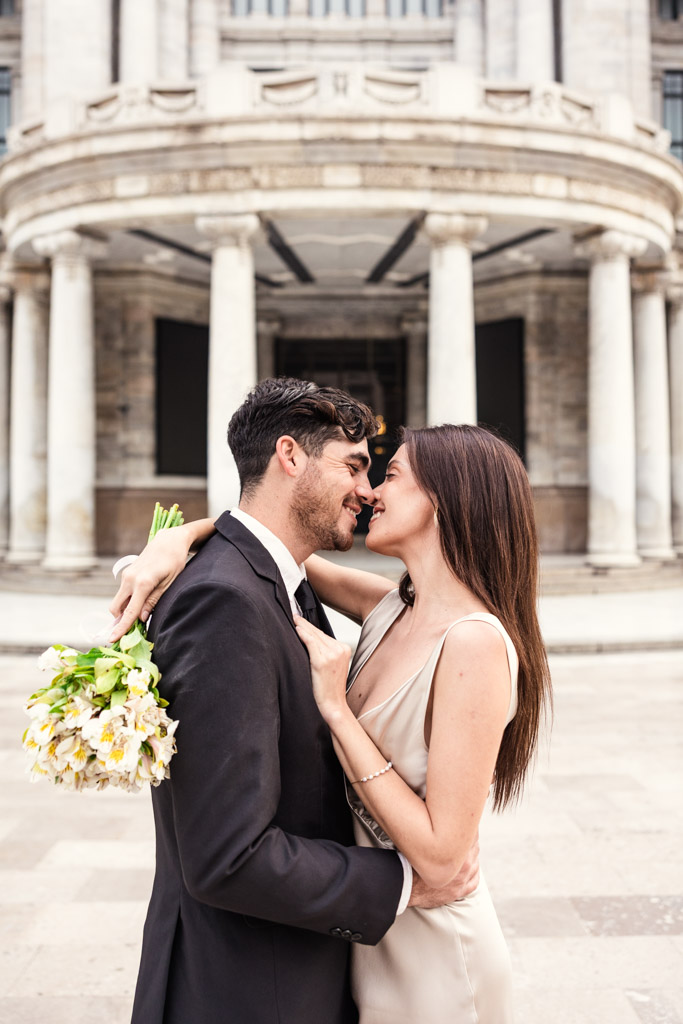 A couple in formal attire embraces and smiles, the bride holding flowers, in front of Palacio de Bellas Artes during their Mexico City elopement