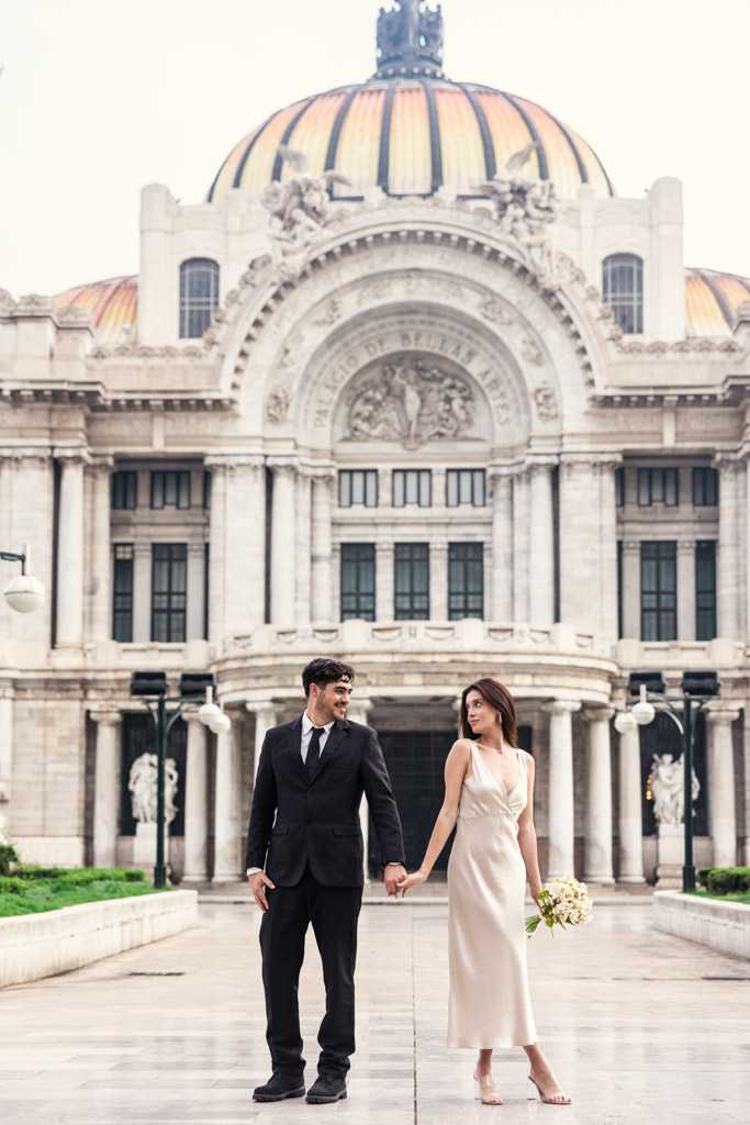 Couple in formal attire holding hands in front of Palacio de Bellas Artes's domed roof during their Mexico City elopement