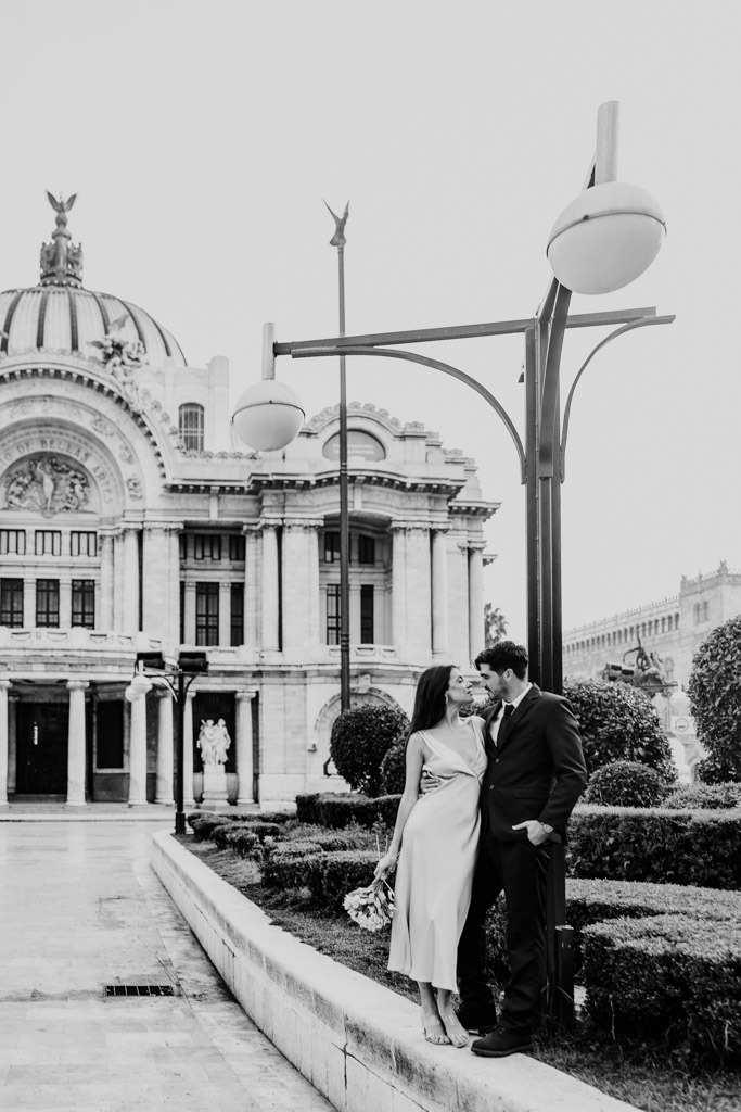 Black and white photo of couple standing on a stone ledge in front of the dome and ornate facade of Palacio de Bellas Artes during their Mexico City elopement