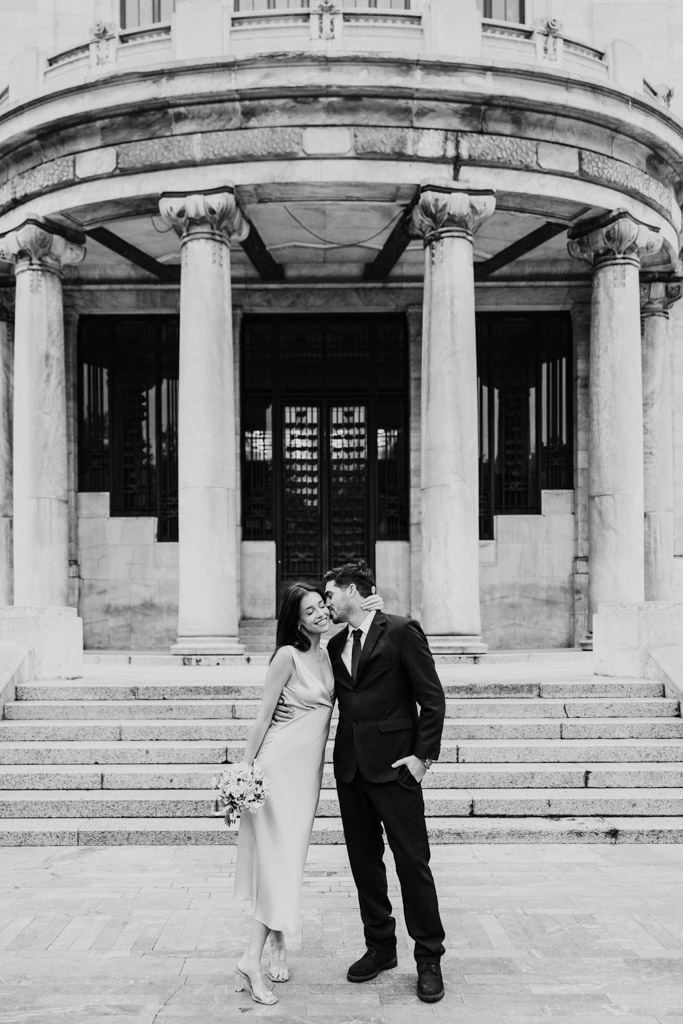 Black and white photo of groom kissing his partner's cheek in front of Palacio de Bellas Artes during their Mexico City elopement.