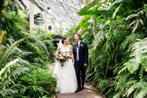A bride and groom smile at each other while walking through the lush, green gardens of The Arbory Chicago.