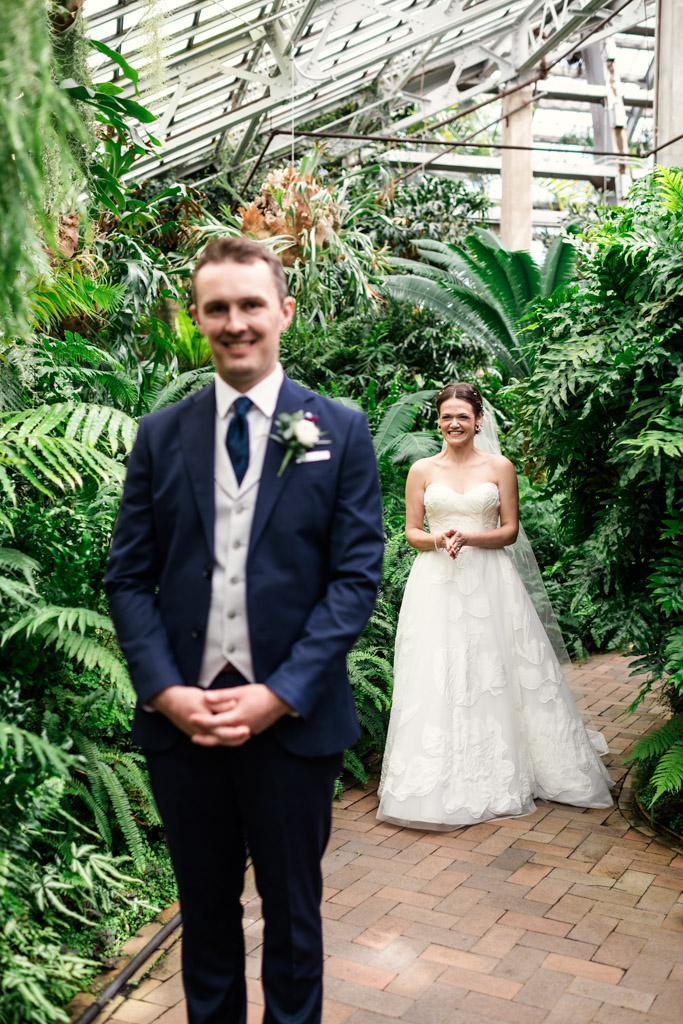 Bride in a white gown smiles as she walks toward the groom in a suit, surrounded by lush greenery in the Fern Room at Garfield Park Conservatory