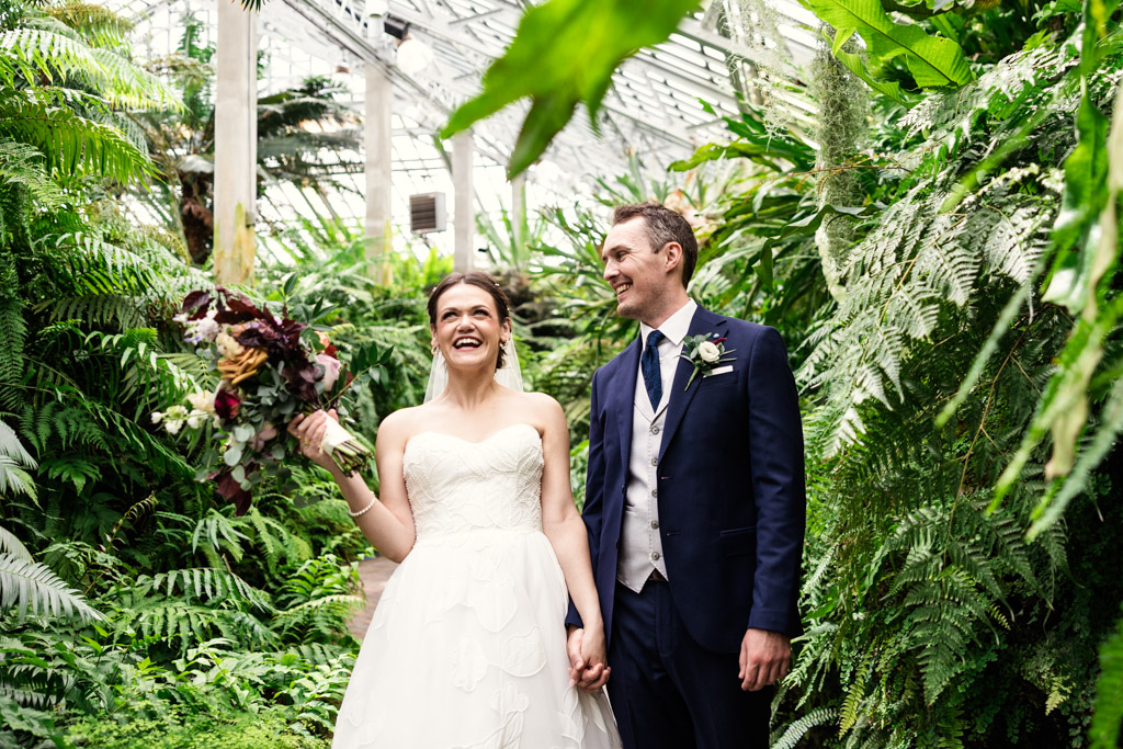 Candid photo of bride and groom holding hands and smiling, surrounded by lush green plants in the Fern Room at Garfield Park Conservatory in Chicago