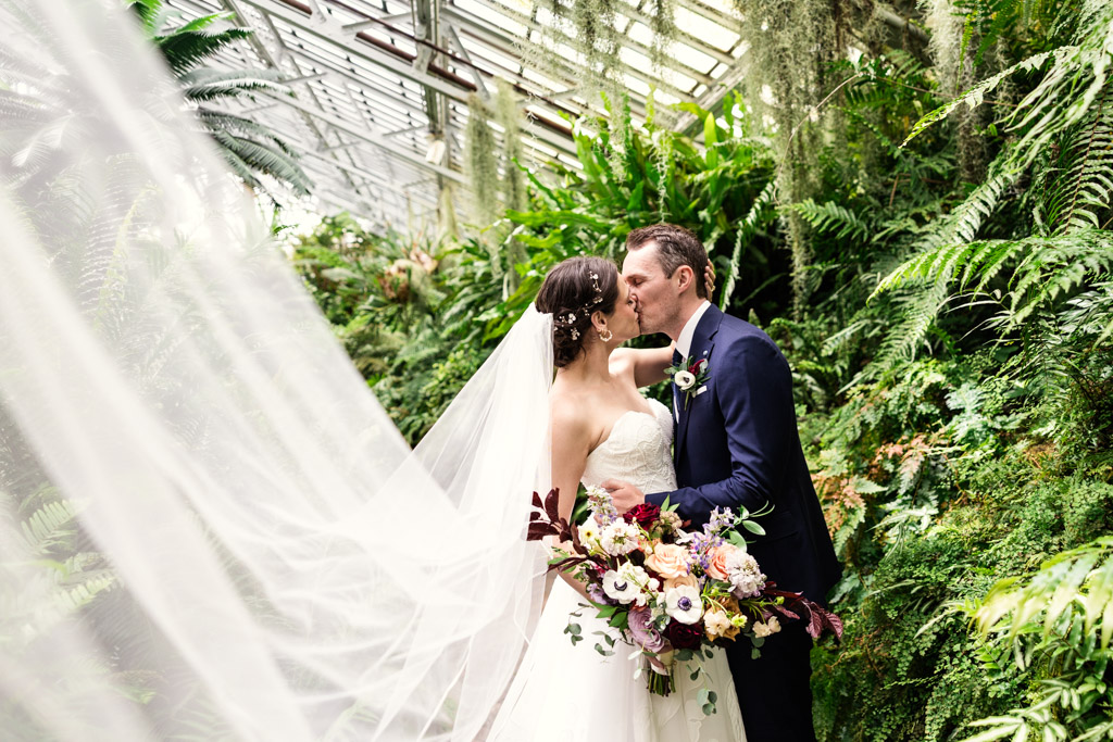 Bride and groom kiss in the Fern Room at Garfield Park Conservatory, surrounded by greenery and the bride’s flowing veil