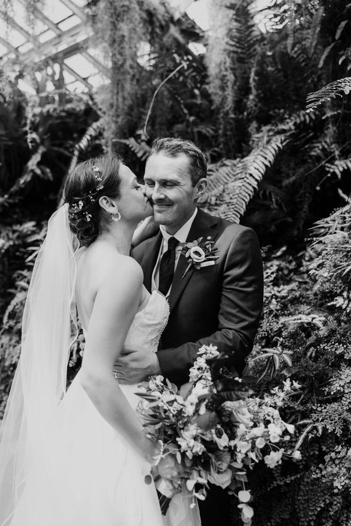 Black and white photo of bride kissing groom on the cheek in the Fern Room at Garfield Park Conservatory