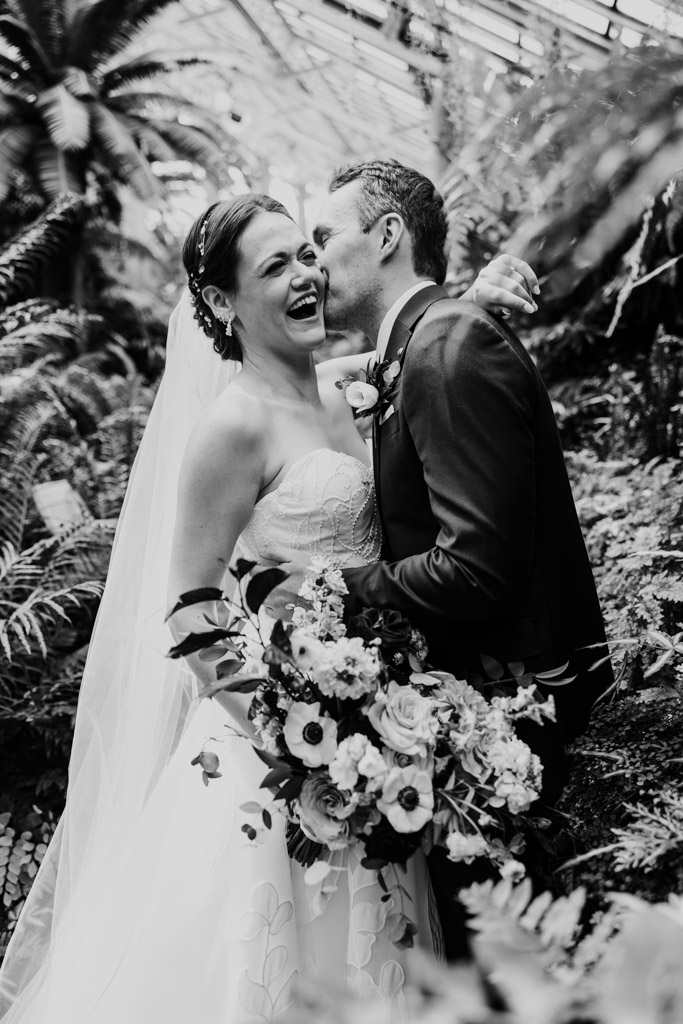 Black and white photo of groom kissing his laughing bride on the cheek inside Garfield Park Conservatory
