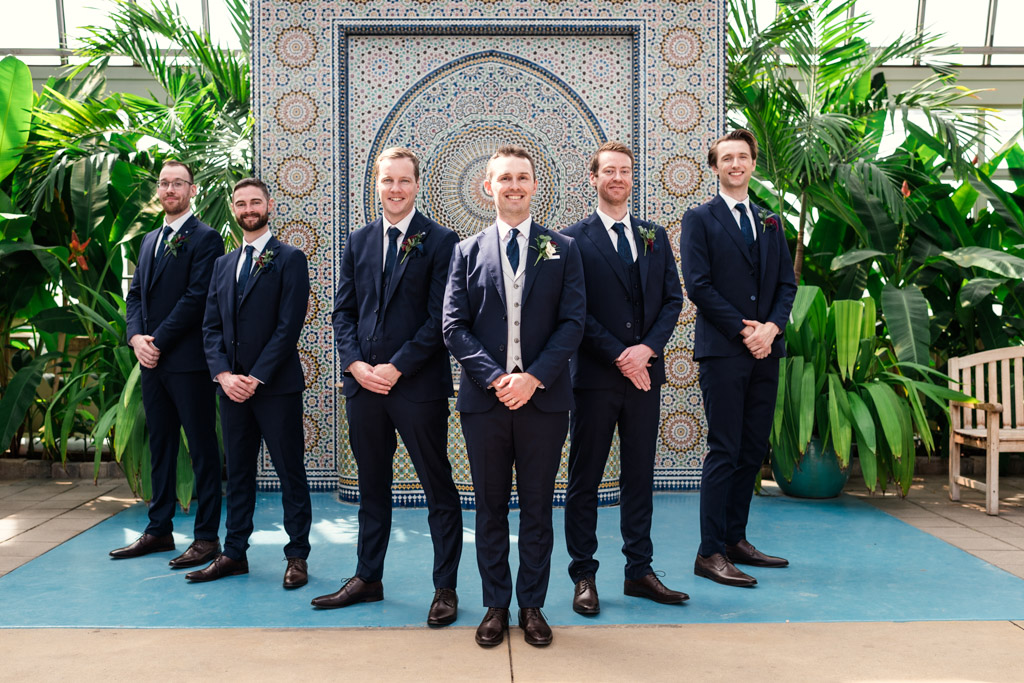 Portrait of groom and groomsmen in front of a decorative tiled wall and lush green plants at Garfield Park Conservatory