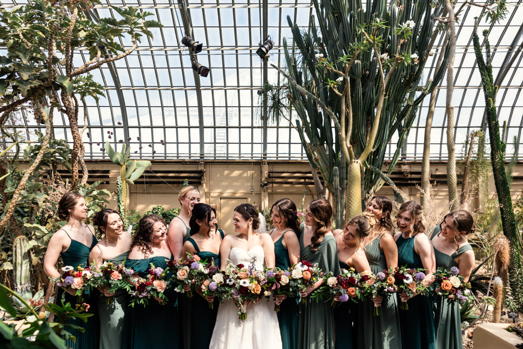 Candid photo of bride in a white dress with bridesmaids in green dresses, holding bouquets in the Desert Room of Garfield Park Conservatory