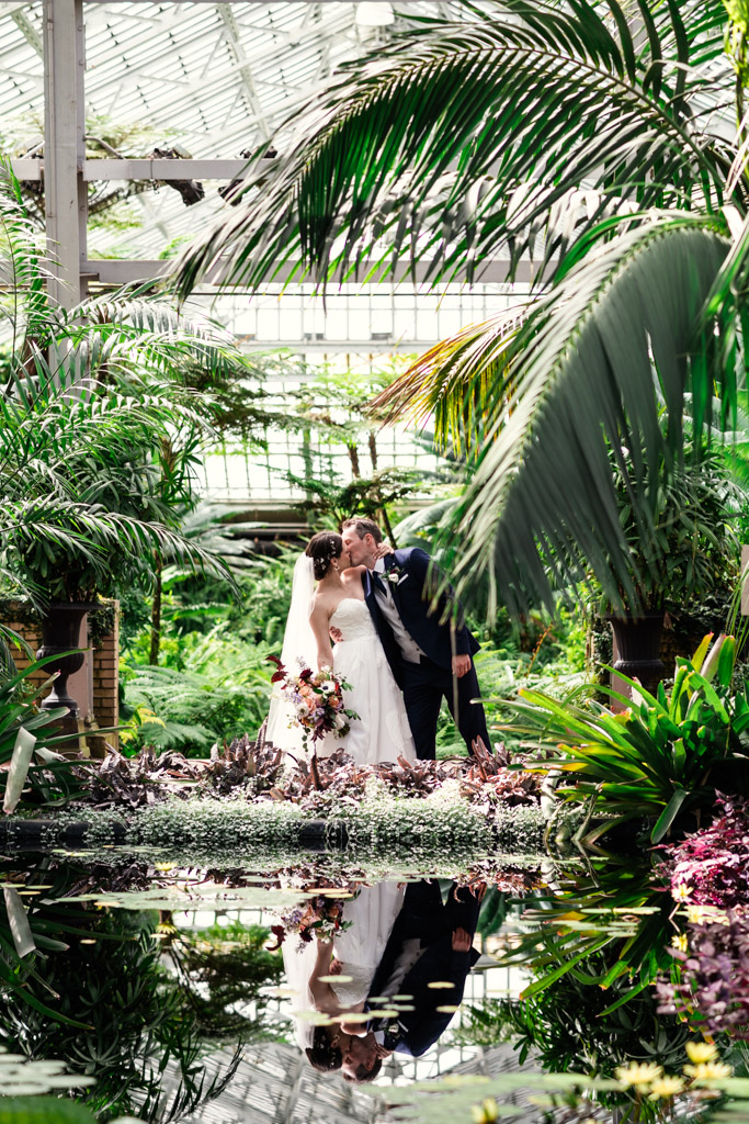 Bride and groom kiss in a lush greenhouse at Garfield Park Conservatory, surrounded by vibrant greenery and reflected in a tranquil pond