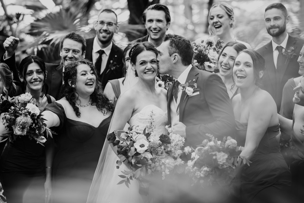 Black and white photo of groom kissing the bride’s cheek as their wedding party celebrates around them at Garfield Park Conservatory