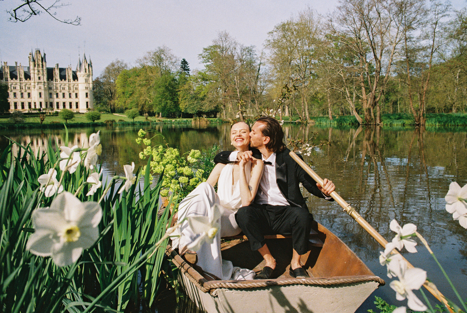 Bride and groom sit in rowboat outside of French castle wedding venue Chateau Challain surrounded by daffodils after their destination wedding ceremony