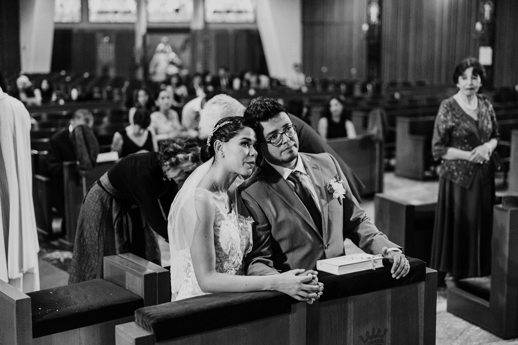 Black and white photo of bride and groom leaning into each other as they kneel at the altar during their wedding ceremony inside Parroquia San Cayetano with guests seated behind them