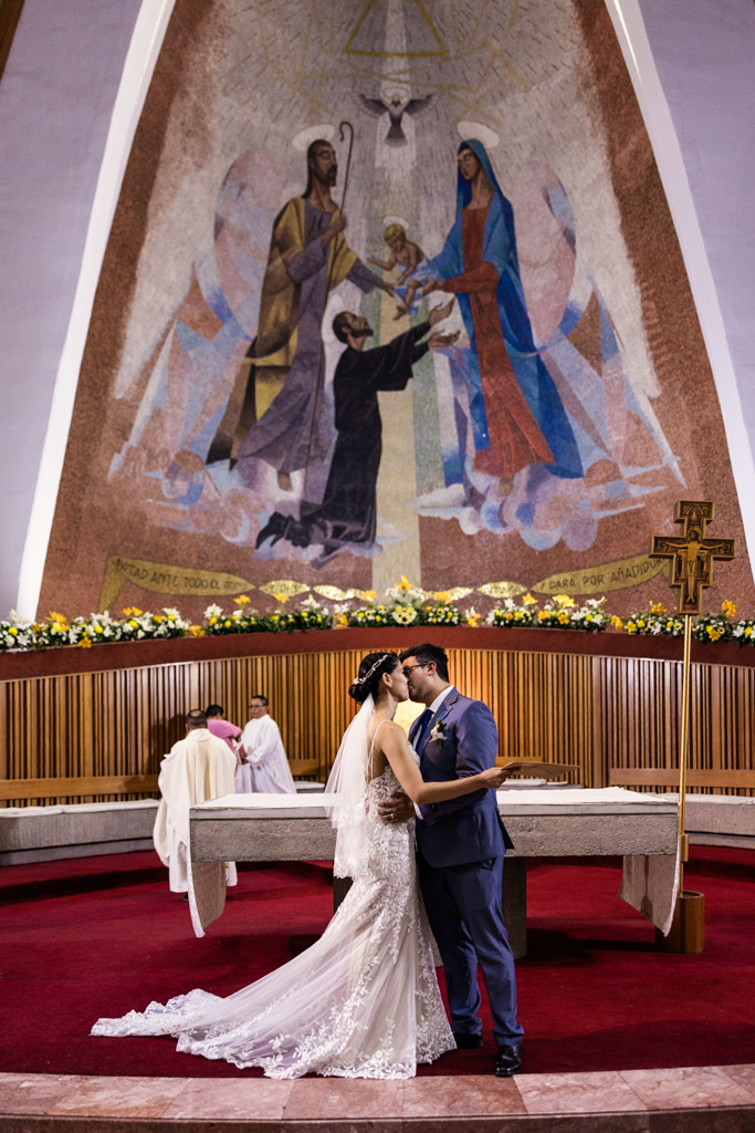 Bride and groom kiss at the altar of Parroquia San Cayetano during their Mexico City wedding ceremony with a large mural behind them