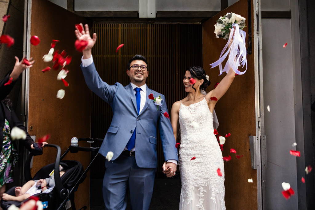 A joyful bride and groom exit Parroquia San Cayetano after their wedding ceremony, smiling and waving as rose petals are thrown around them