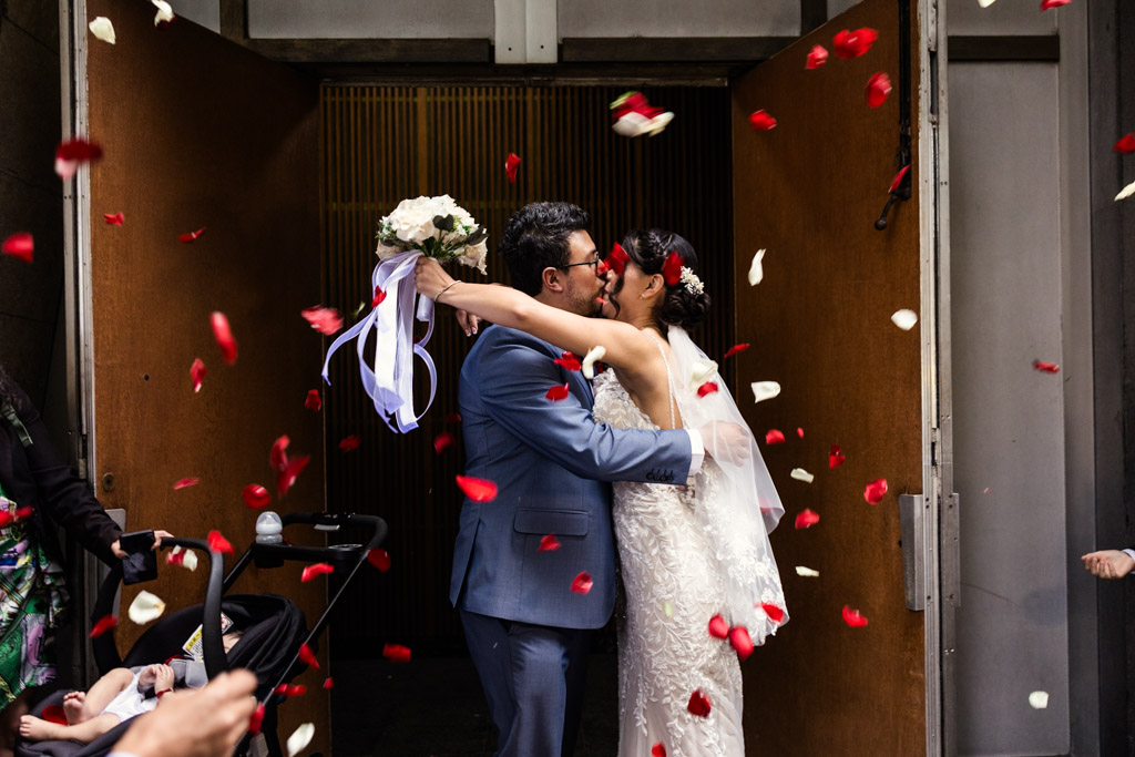 Bride and groom kiss surrounded by falling red rose petals outside Parroquia San Cayetano after their wedding ceremony