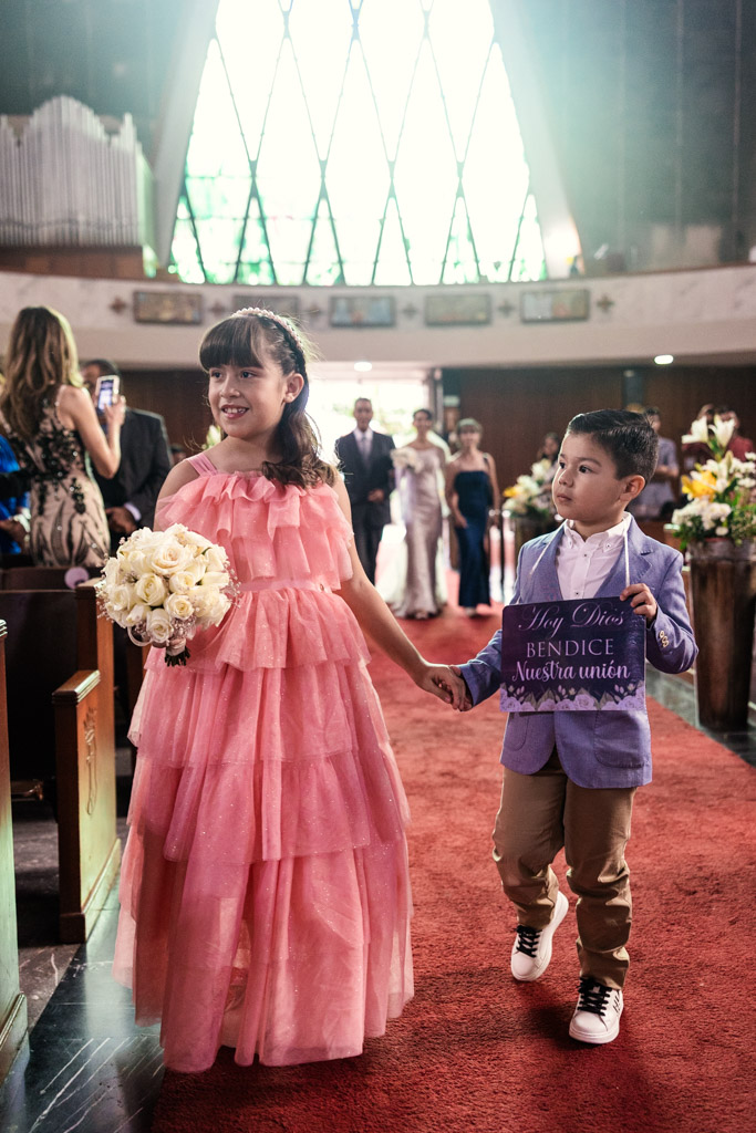 Flower girl in a pink dress and a ring bearer in a suit walk down the aisle holding hands during wedding ceremony at Parroquia San Cayetano in Mexico City