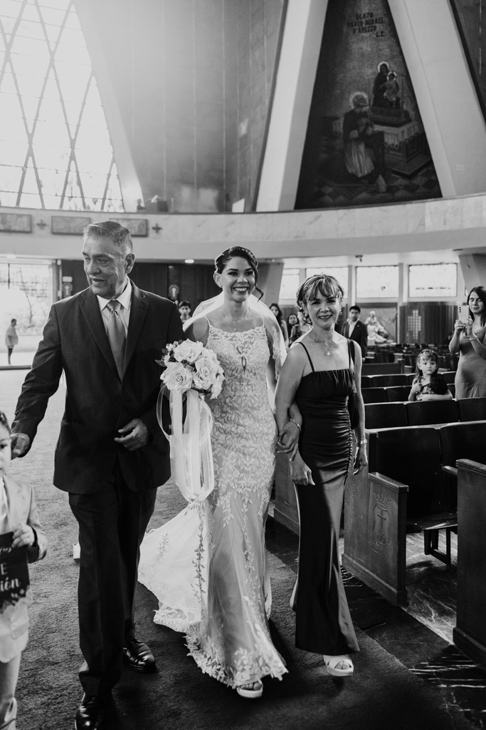 Black and white photo of bride in a lace dress walking down the aisle with her parents inside Parroquia San Cayetano