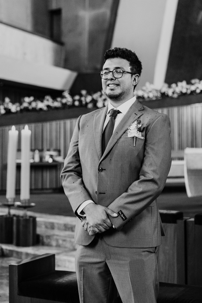 Black and white photo of happy groom in a suit watching bride wlak down the aisle during his Parroquia San Cayetano wedding cermony , hands clasped, with candles and flowers in the background
