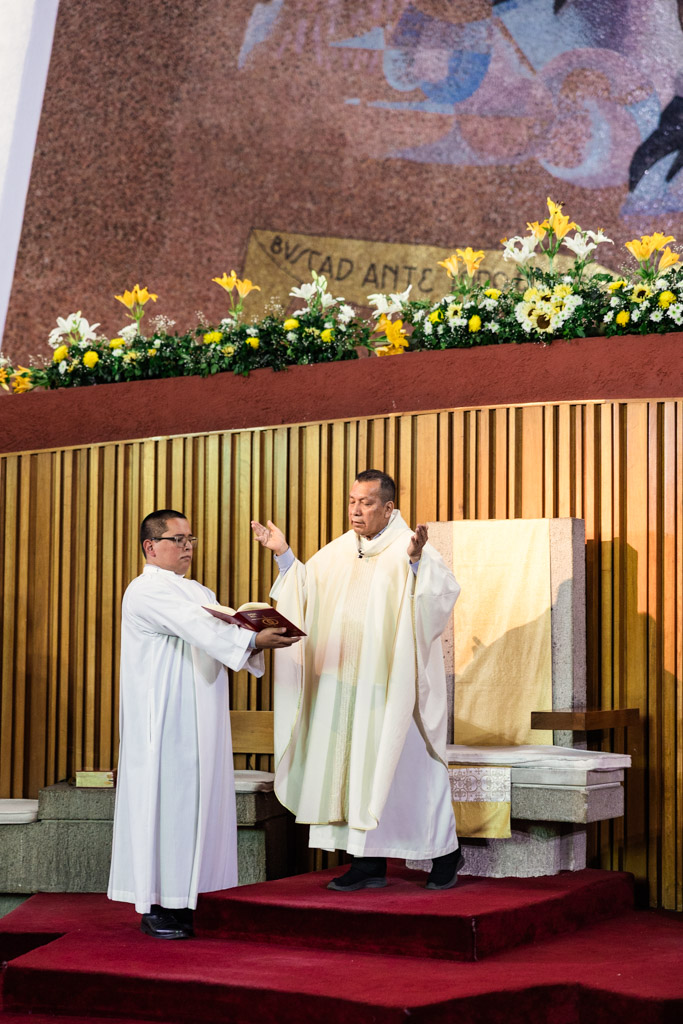 Two priests in white robes stand at an altar during a Mexico City wedding church service, with flowers and a mural in the background.