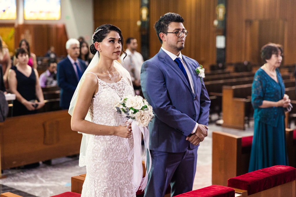 Bride and groom stand together inside Parroquia San Cayetano during their wedding ceremony, the bride holding flowers, with guests seated in the background