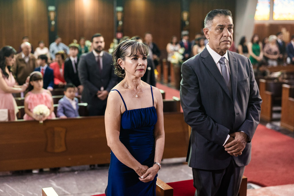 Bride's mother in a blue dress and father in a suit stand solemnly in Parroquia San Cayetano during wedding ceremony, with guests seated behind them