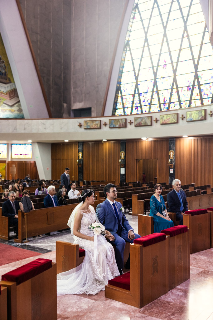 Bride and groom seated at the altar inside Parroquia San Cayetano during their wedding ceremony, with guests in pews and vibrant stained glass windows behind them