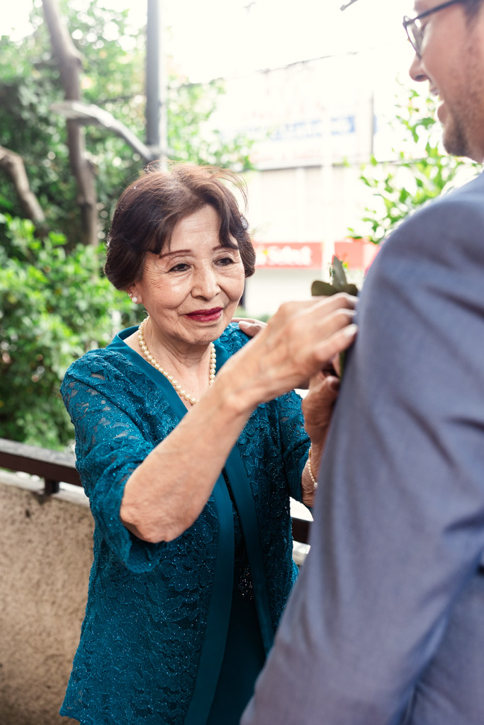 An elderly woman pins a boutonniere on a man's suit jacket outdoors at a Mexico City wedding, both smiling.