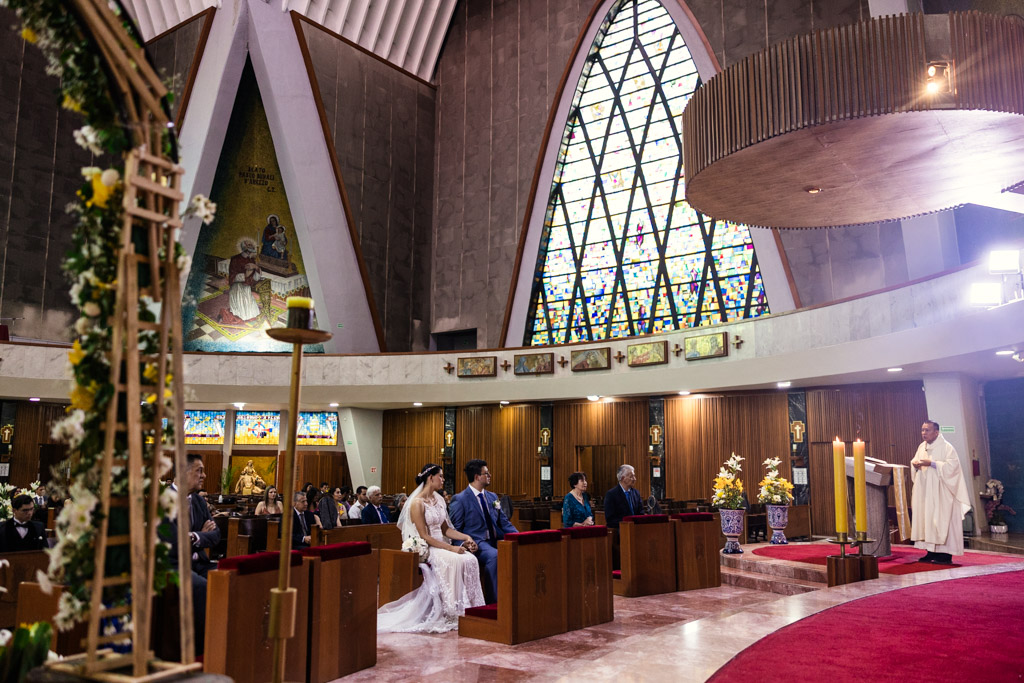 Bride and groom seated with guests inside Parroquia San Cayetano during their wedding ceremony, the priest speaking from the lectern
